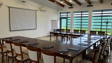 Une salle de conférence équipée d’une longue table, de chaises, d’un écran de projection et de grandes fenêtres donnant sur un champ verdoyant.