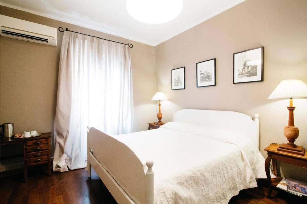 A hotel room with a bed, a nightstand, a lamp, a desk with a lamp, and a window with curtains.