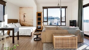 Moderni hotellihuone, jossa on sänky, sohva, pieni pöytä ja suuri ikkuna, josta avautuu näkymä kaupunkiin.