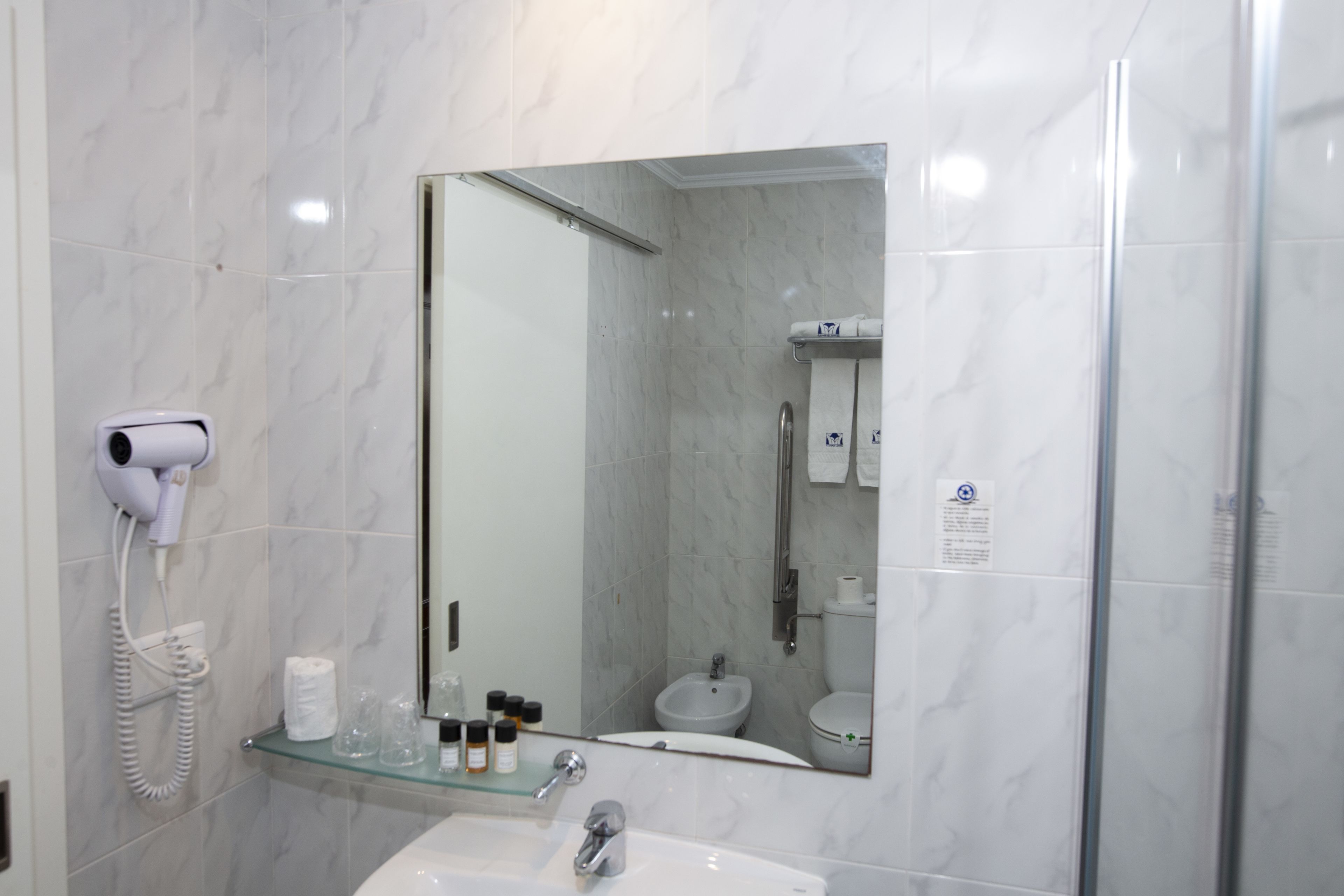 A bathroom with a glass shelf holding toiletries, a hairdryer, and a towel dispenser.