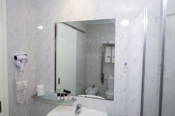 Une salle de bain avec une étagère en verre où sont rangés les articles de toilette, un sèche-cheveux et un distributeur de serviettes.