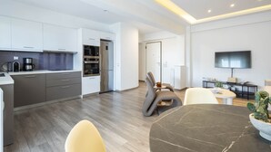 A modern kitchen with white cabinets, a built-in oven, and a coffee maker. There is a dining area with a round table and chairs, and a flat-screen TV mounted on the wall.