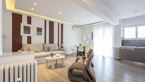 A modern living room with a sofa, coffee table, and unique chair, a dining area with chairs, and a kitchen with cabinets.