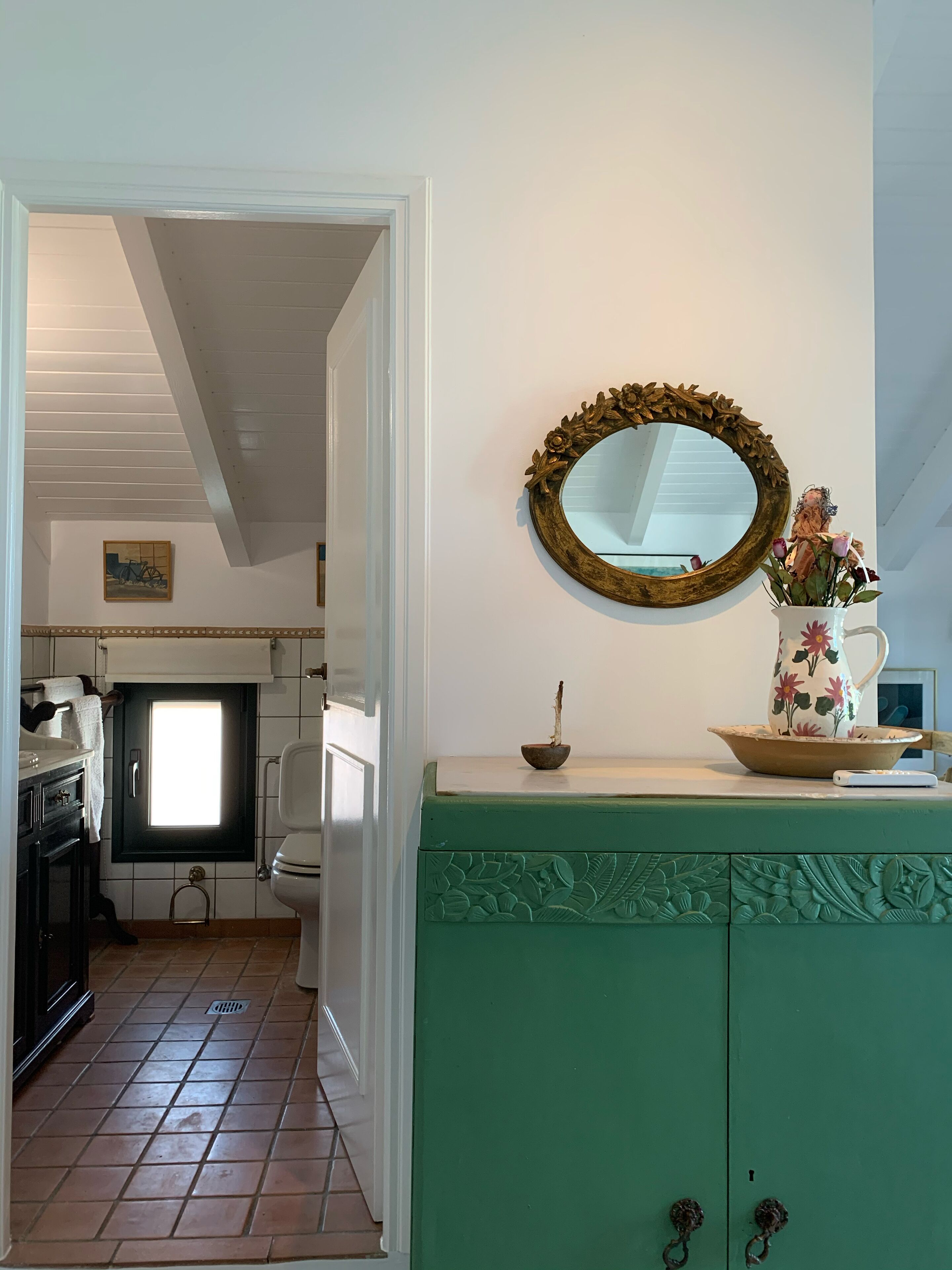 A bathroom with a green cabinet, a mirror with a floral frame, and a small window.
