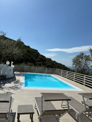 A swimming pool with a white fence, lounge chairs, and a view of a mountainous landscape.