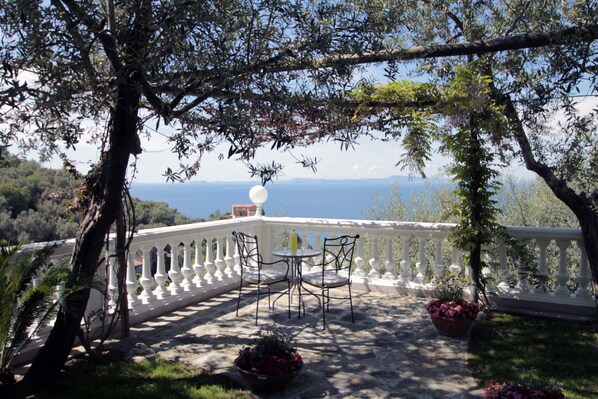 A terrace with a white balustrade, two tables, and chairs, surrounded by potted plants and overlooking a scenic view.