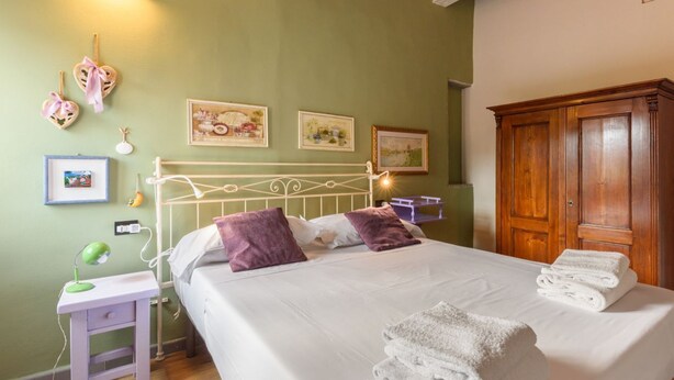 A neatly made bed with white linens and purple pillows, a bedside table with a lamp, and a wooden wardrobe. - Apartment Cappelle Medicee Florence by Mmega (Firenze)