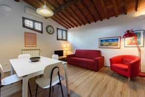 Un coin repas avec une table et des chaises blanches, un canapé rouge et un fauteuil rouge. On y trouve des fenêtres, une horloge et une lampe fixée au mur.