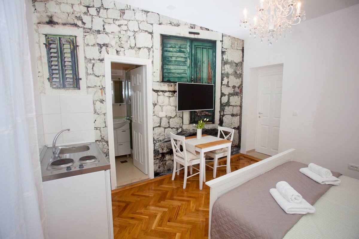A compact room with a bed, a small table with chairs, a TV, and a kitchenette with a sink and stove.