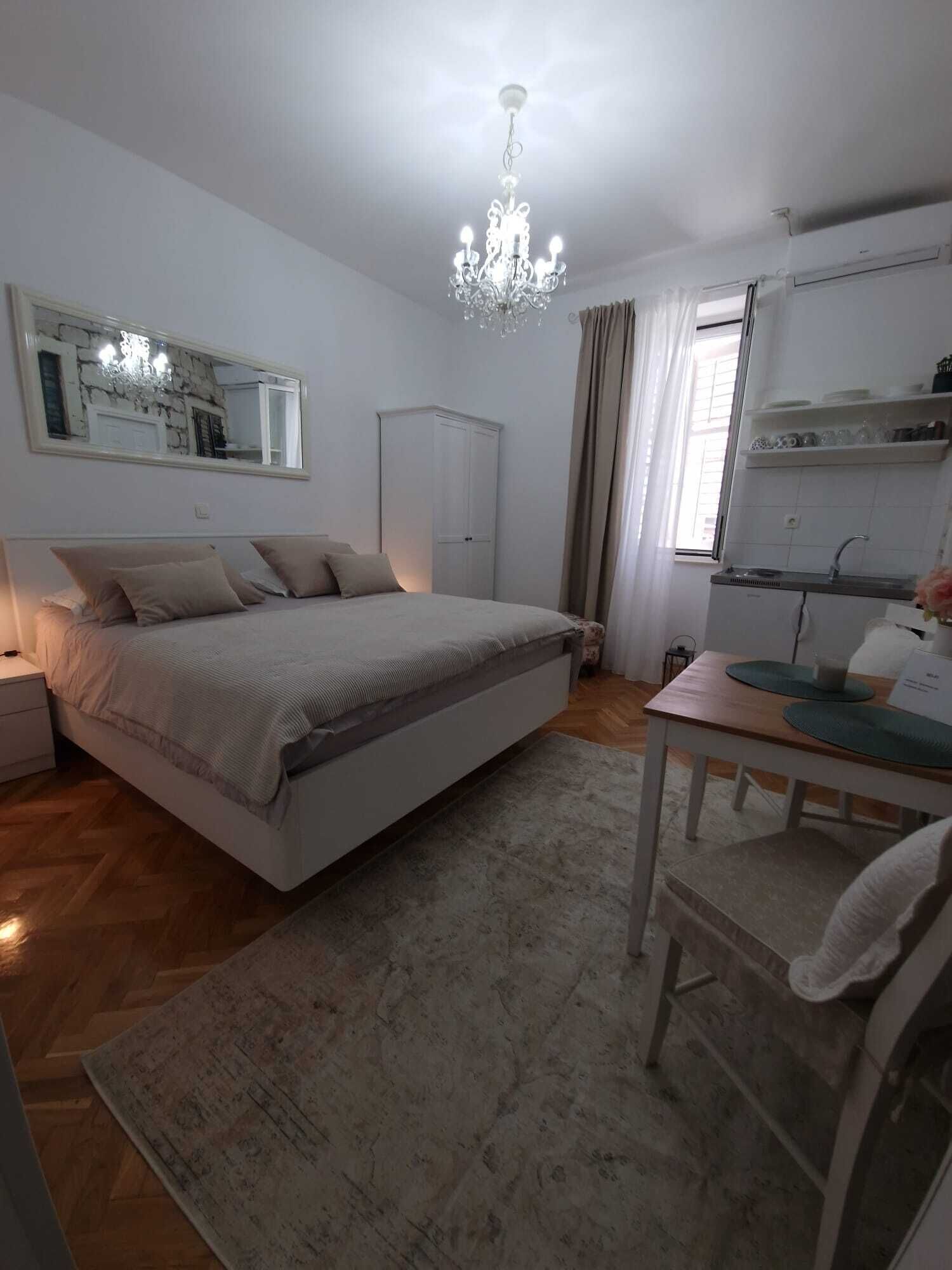 A bedroom with a bed, a nightstand, a desk with a chair, a chandelier, a wardrobe, and a window with curtains.
