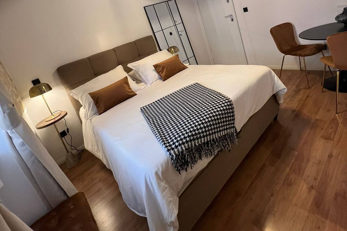 A modern hotel room with a large bed, a bedside table with a lamp, a small round table with chairs, and a ceiling light fixture.