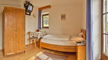 Un dormitorio con una cama de madera, una mesita con una silla, un televisor montado en la pared y una ventana con vista a un paisaje verde.