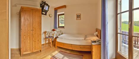 A bedroom with a wooden bed, a small table with a chair, a TV mounted on the wall, and a window with a view of a green landscape.