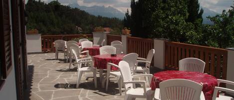 An outdoor patio with stone flooring, white plastic chairs, and red tablecloths.
