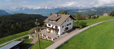 A two-story house with a balcony, surrounded by green fields and mountains in the background.