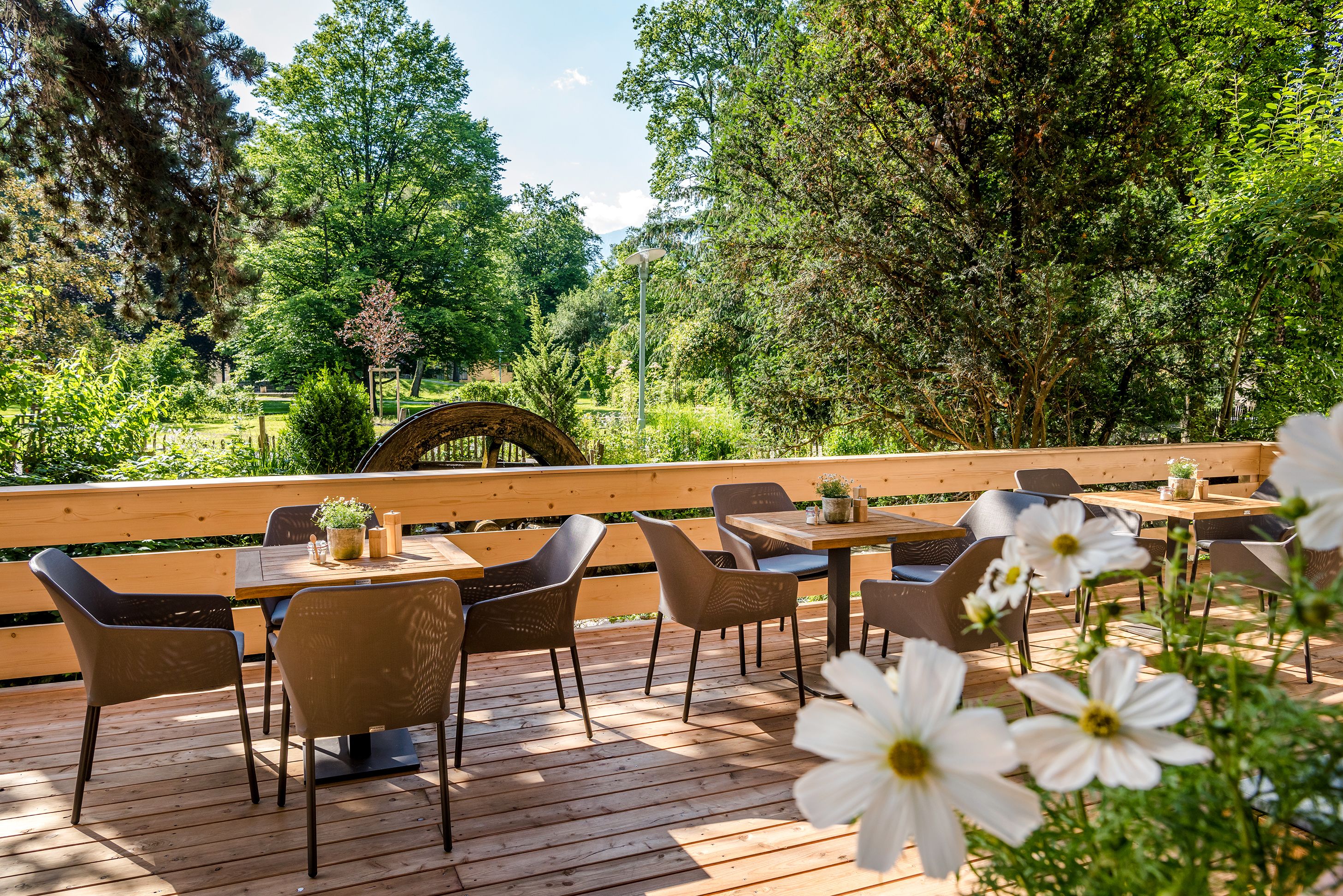 An outdoor patio with wooden tables and chairs, surrounded by greenery and trees.