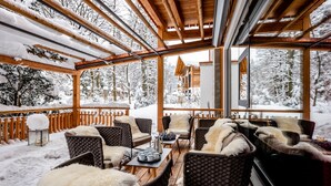 A wooden deck with wicker furniture and fur throws, overlooking a snowy forest and a cabin. - Werdenfelserei (Garmisch-Partenkirchen)