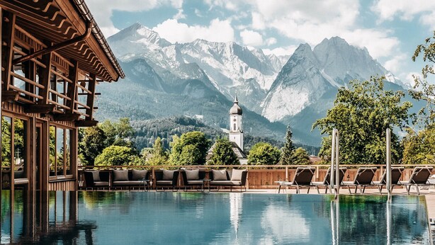A pool with lounge chairs, a wooden building, and a church with a bell tower, surrounded by mountains. - Werdenfelserei (Garmisch-Partenkirchen)
