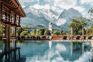 Une piscine avec des chaises longues, un bâtiment en bois et une église dotée d’un clocher, le tout entouré de montagnes.