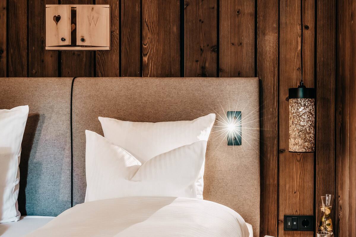 Una cama bien hecha con sábanas blancas, cabecera de madera, mesita de noche con lámpara y un libro.