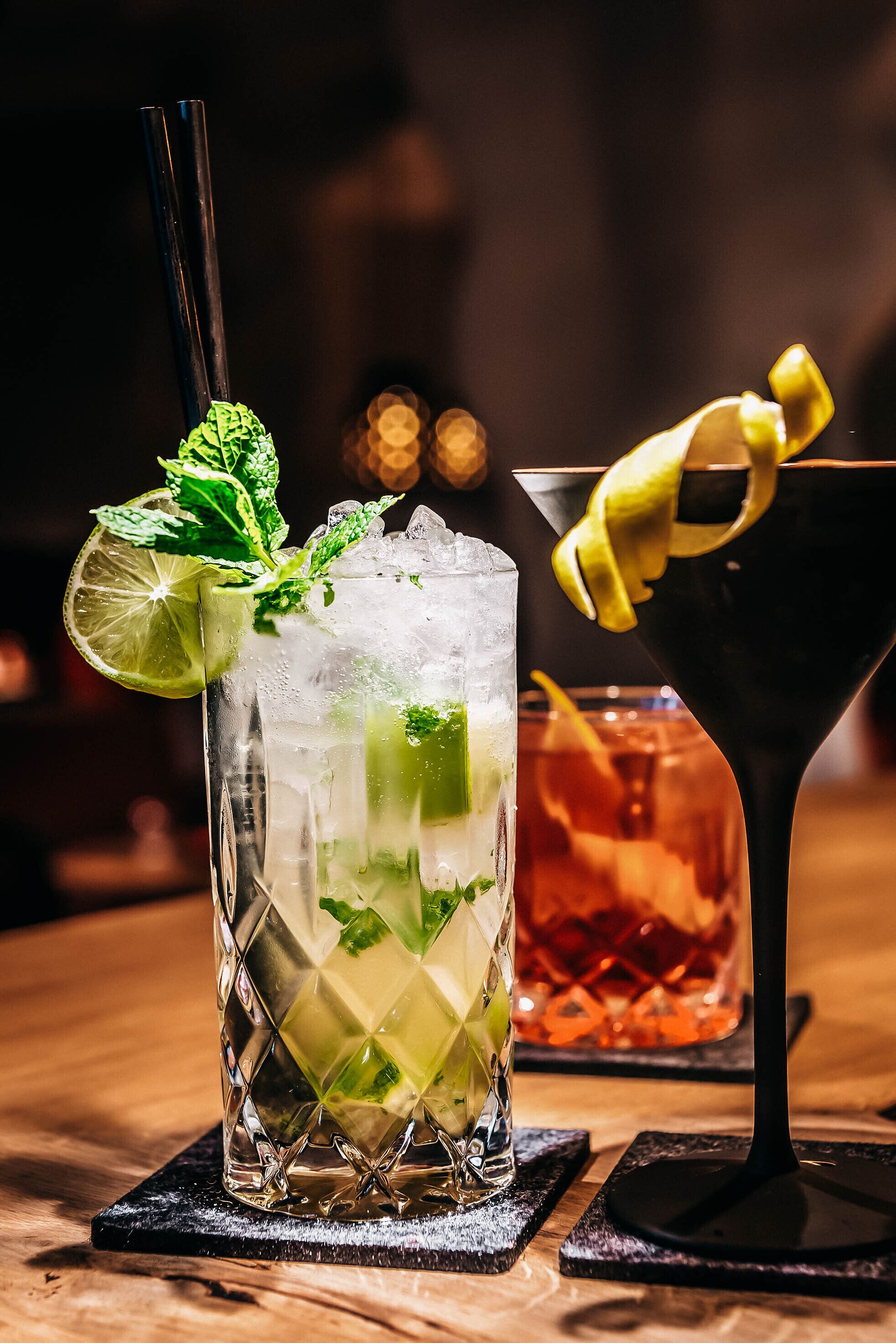 A glass of iced drink garnished with lime and mint, a cocktail in a martini glass, and a dark drink in a glass with a lemon twist.