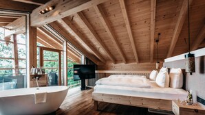 A modern bedroom with a large bathtub, a bed with white bedding, wooden beams, and a TV. - Werdenfelserei (Garmisch-Partenkirchen)
