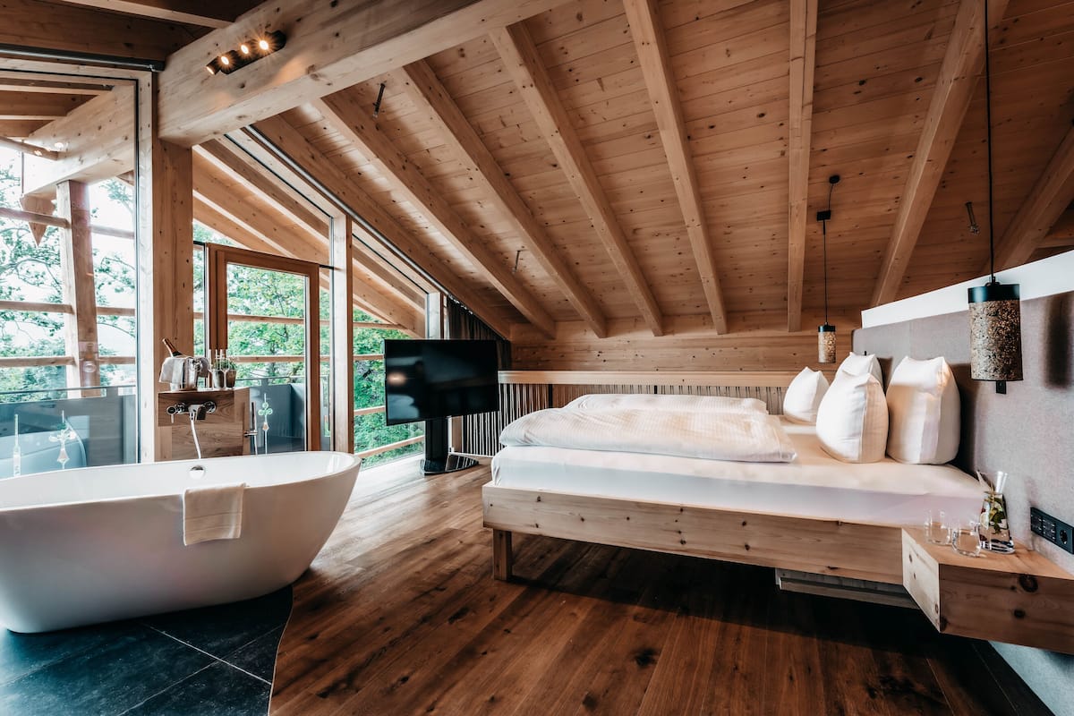 Un dormitorio moderno con una bañera grande, una cama con ropa de cama blanca, vigas de madera y un televisor.