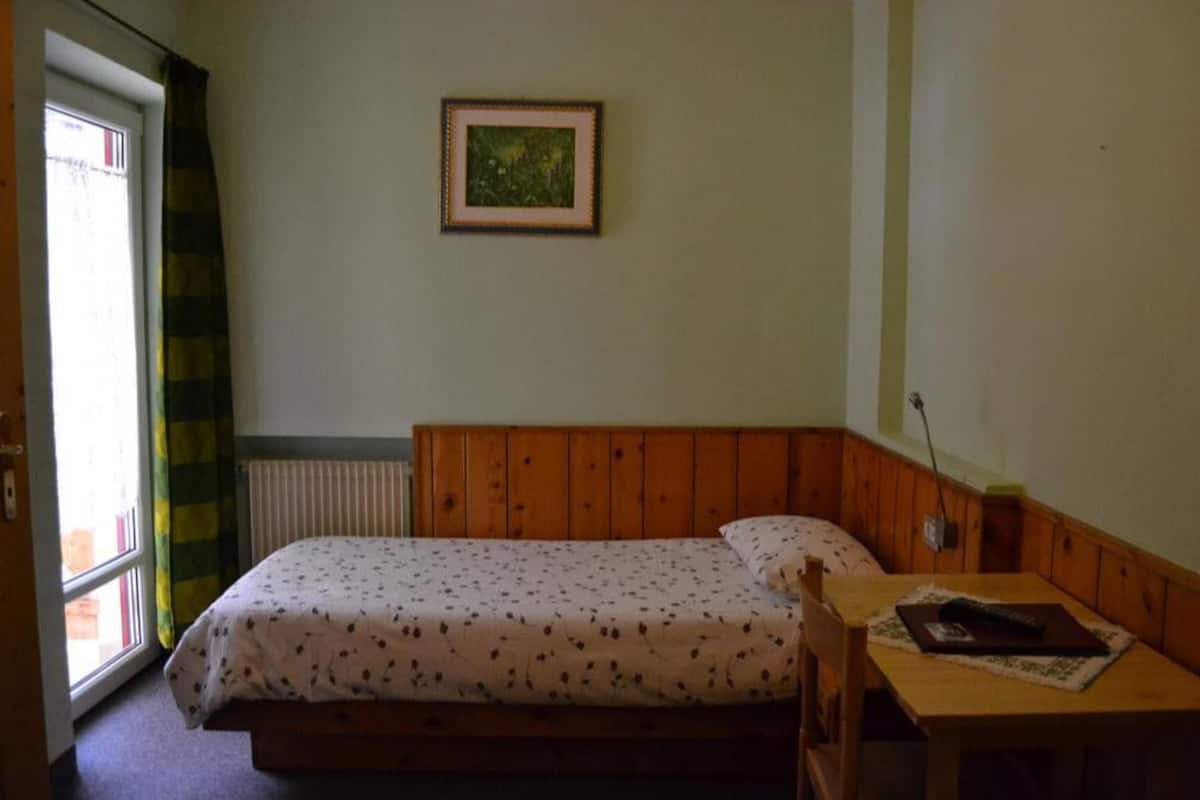 A small, single-bed room with a wooden headboard, a framed picture on the wall, and a window with curtains.