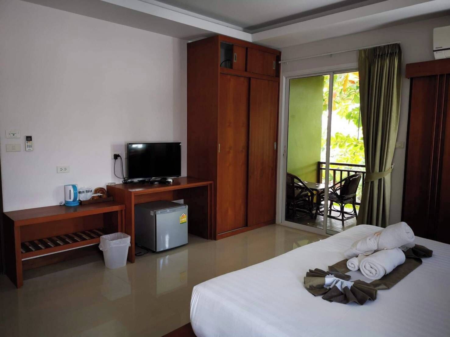 Habitación de hotel con cama, escritorio con televisor, mini refrigerador, bote de basura y balcón con sillas.