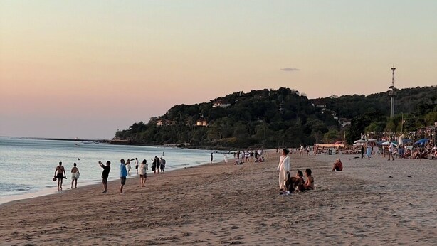 A beach with people walking and sitting, a hill with buildings in the background, and a tower on the hill. - The Peace Tara House (Ko Lanta)