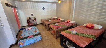 Una habitación con cuatro camas, cada una con un edredón de diseño diferente. Hay dos mesitas con sillas, y un estante con diversos objetos.