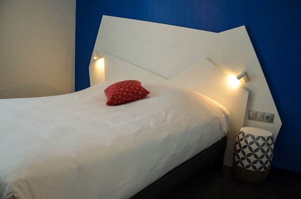 A neatly made bed with white bedding and a red geometric pillow, a bedside lamp, and a patterned ottoman. - Hotel Origami (Strasbourg)
