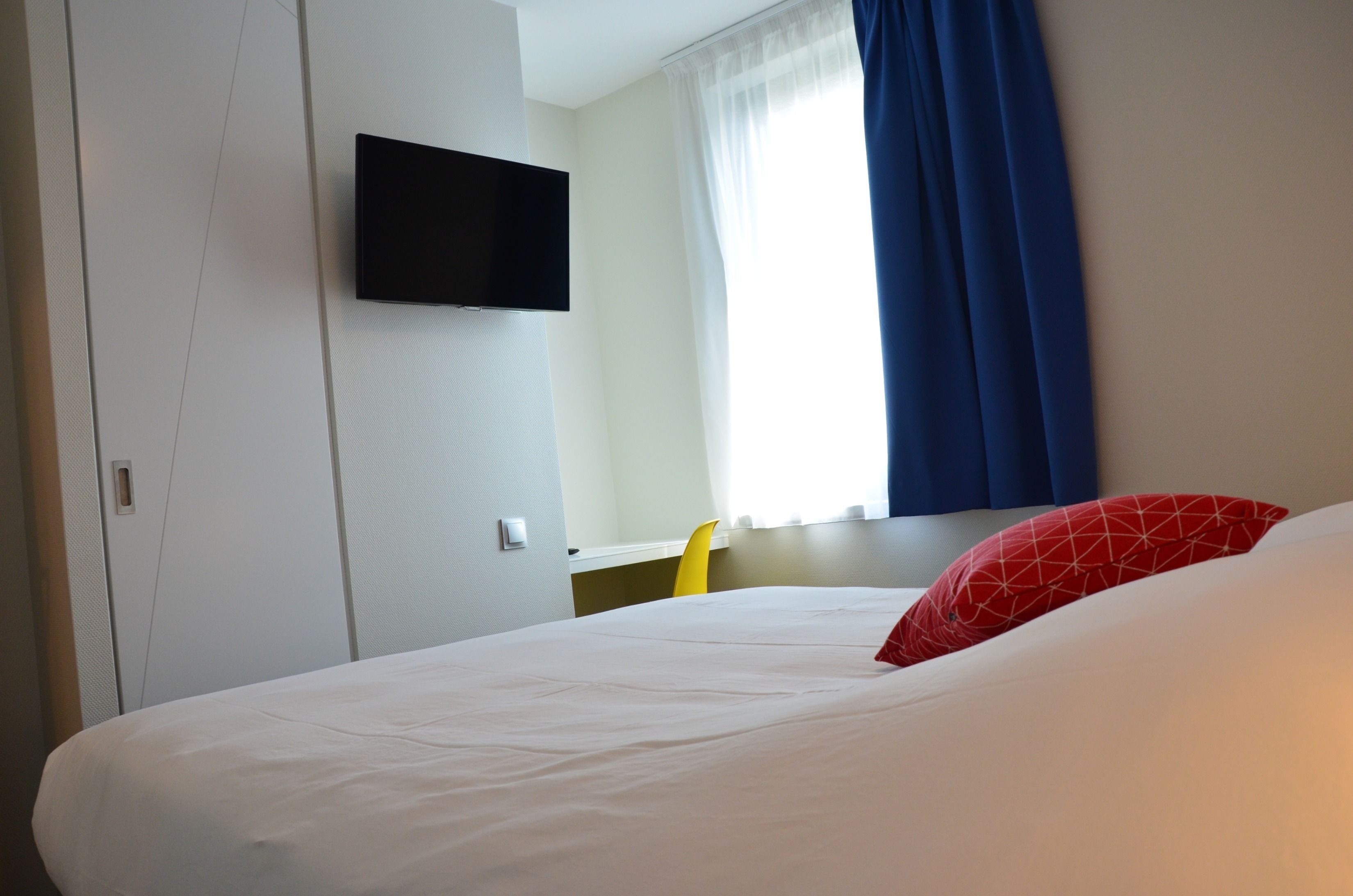 A hotel room with a bed, a TV mounted on the wall, a window with blue curtains, and a yellow chair.