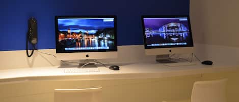 Dos computadoras Apple iMac sobre un escritorio blanco frente a una pared azul, con una silla blanca al frente.