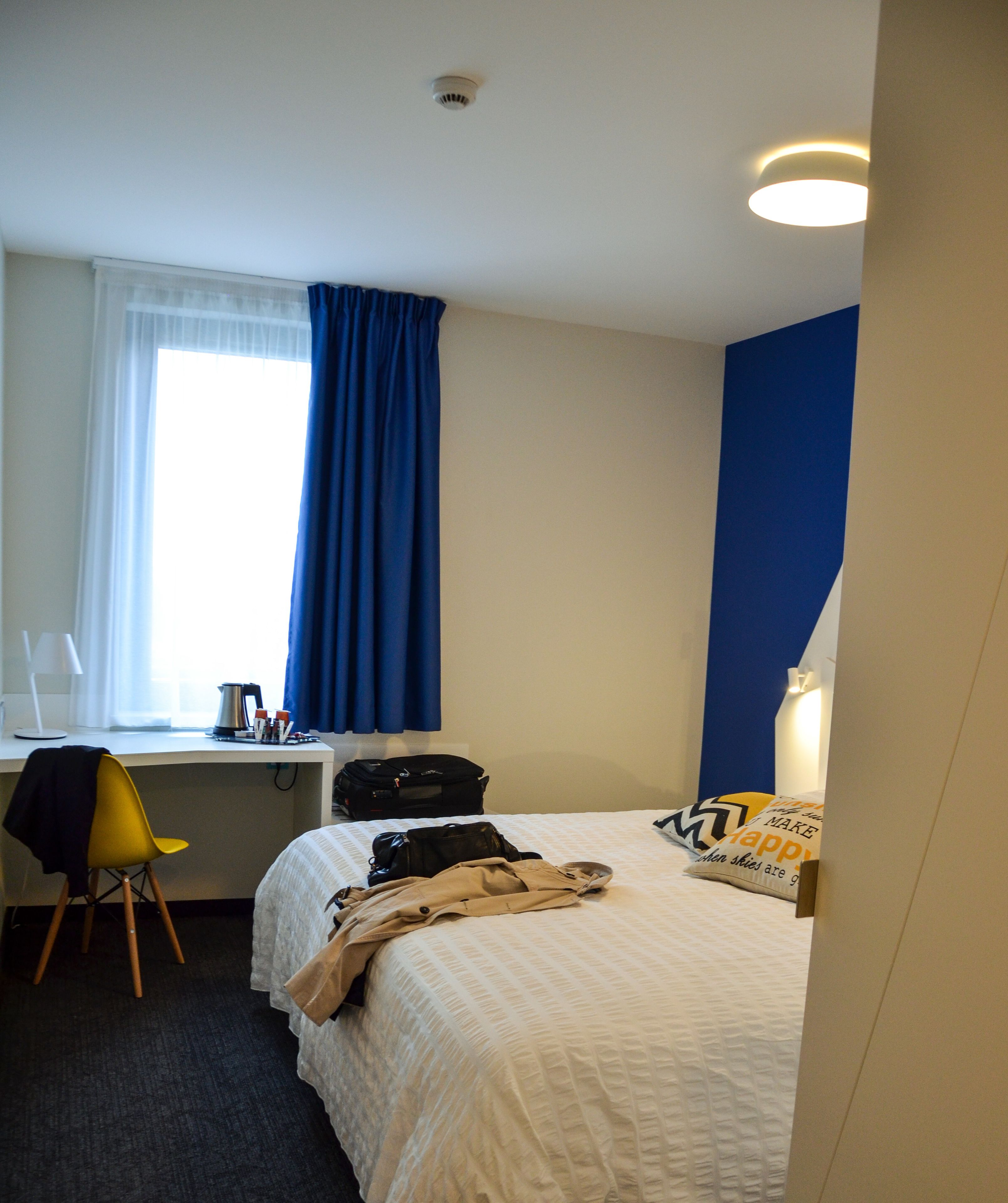 Habitación de hotel con cama, escritorio con silla, ventana con cortinas, mesita de noche y lámpara.