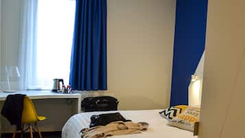 Habitación de hotel con cama, escritorio con silla, ventana con cortinas, mesita de noche y lámpara.