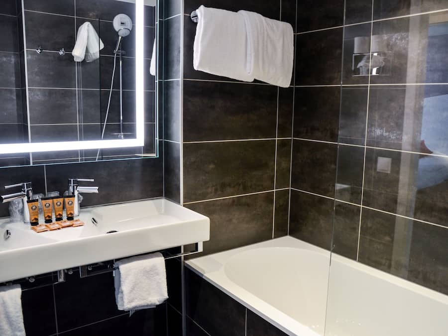 A modern bathroom with a large mirror, a white bathtub, and a sink with toiletries.