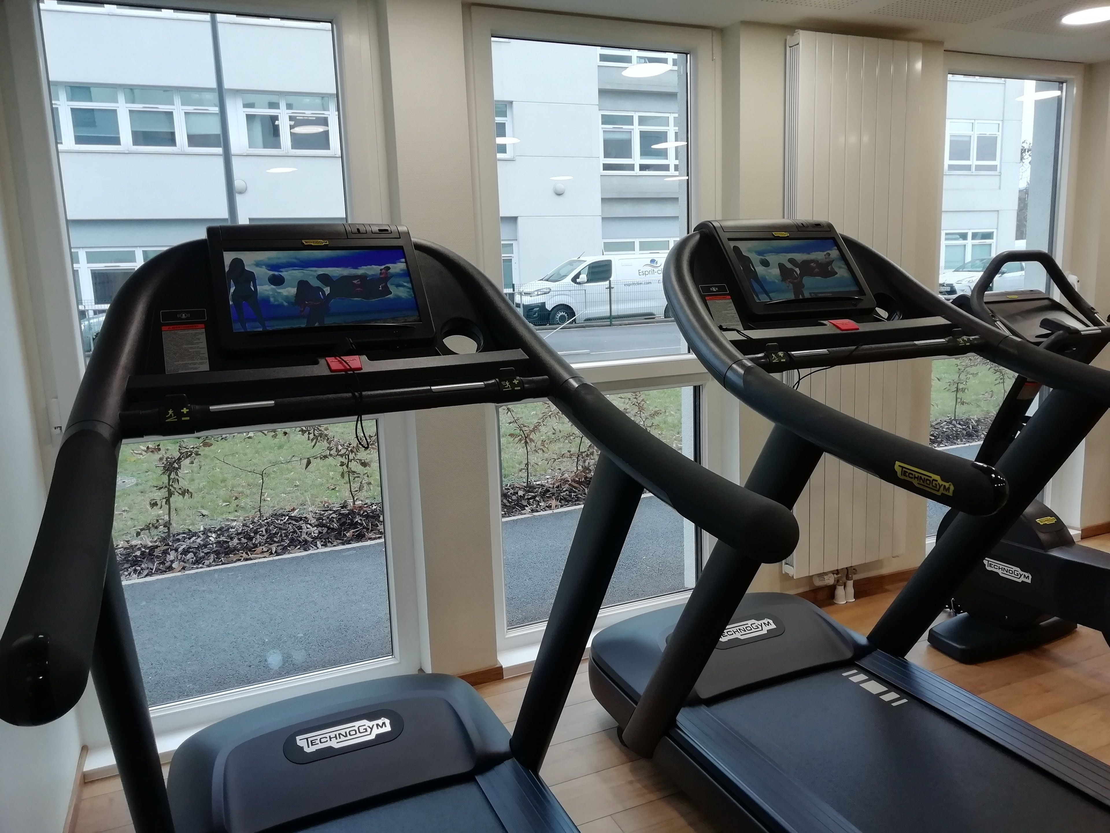 A gym with treadmills equipped with screens, large windows, and a view of a parking lot.