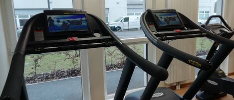 A gym with treadmills equipped with screens, large windows, and a view of a parking lot.
