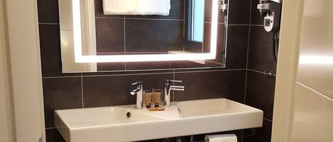 Un baño moderno con un lavamanos blanco, paredes cubiertas de baldosas oscuras y un espejo grande con iluminación integrada.