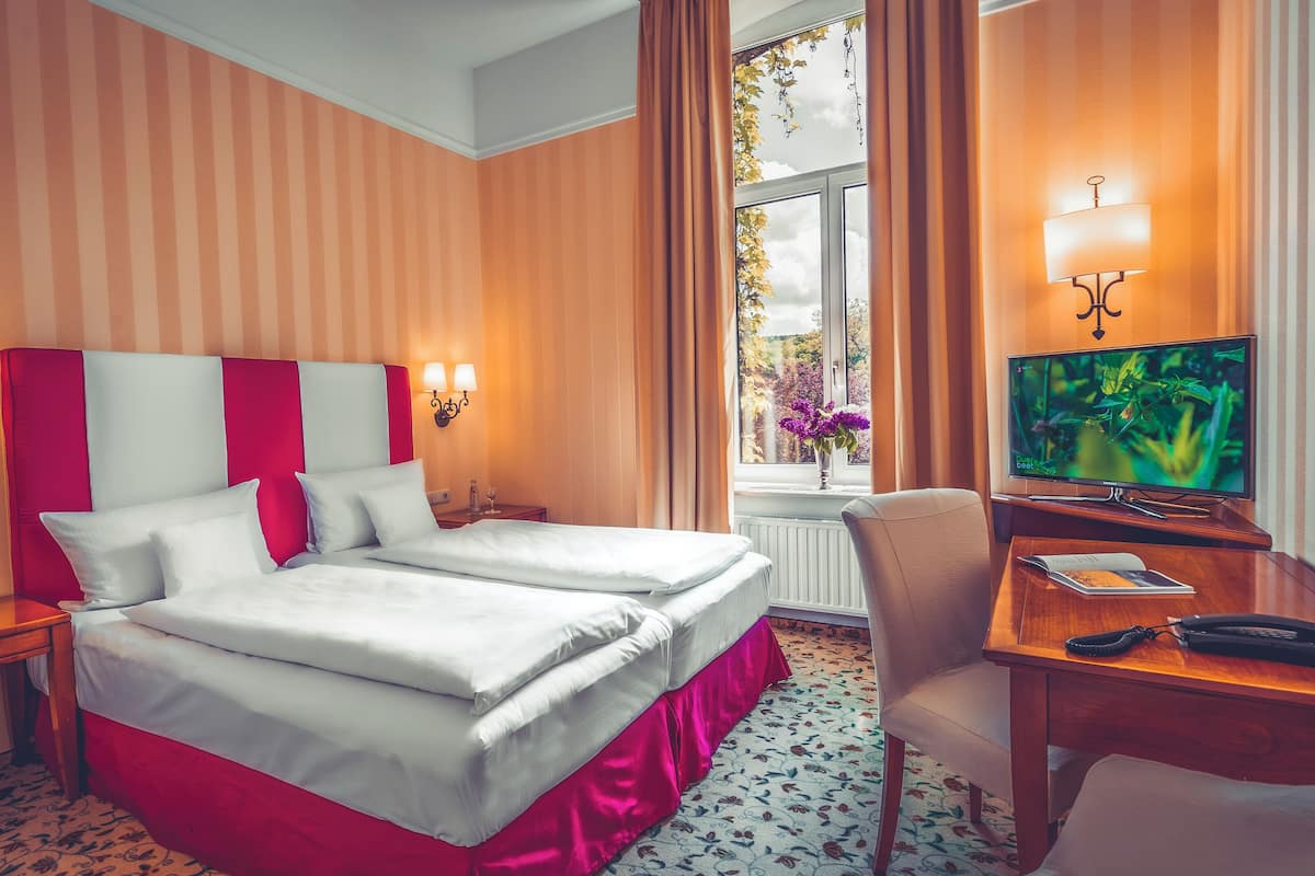 A hotel room with a double bed, two bedside tables with lamps, a television, a desk with a chair, and a window with curtains.