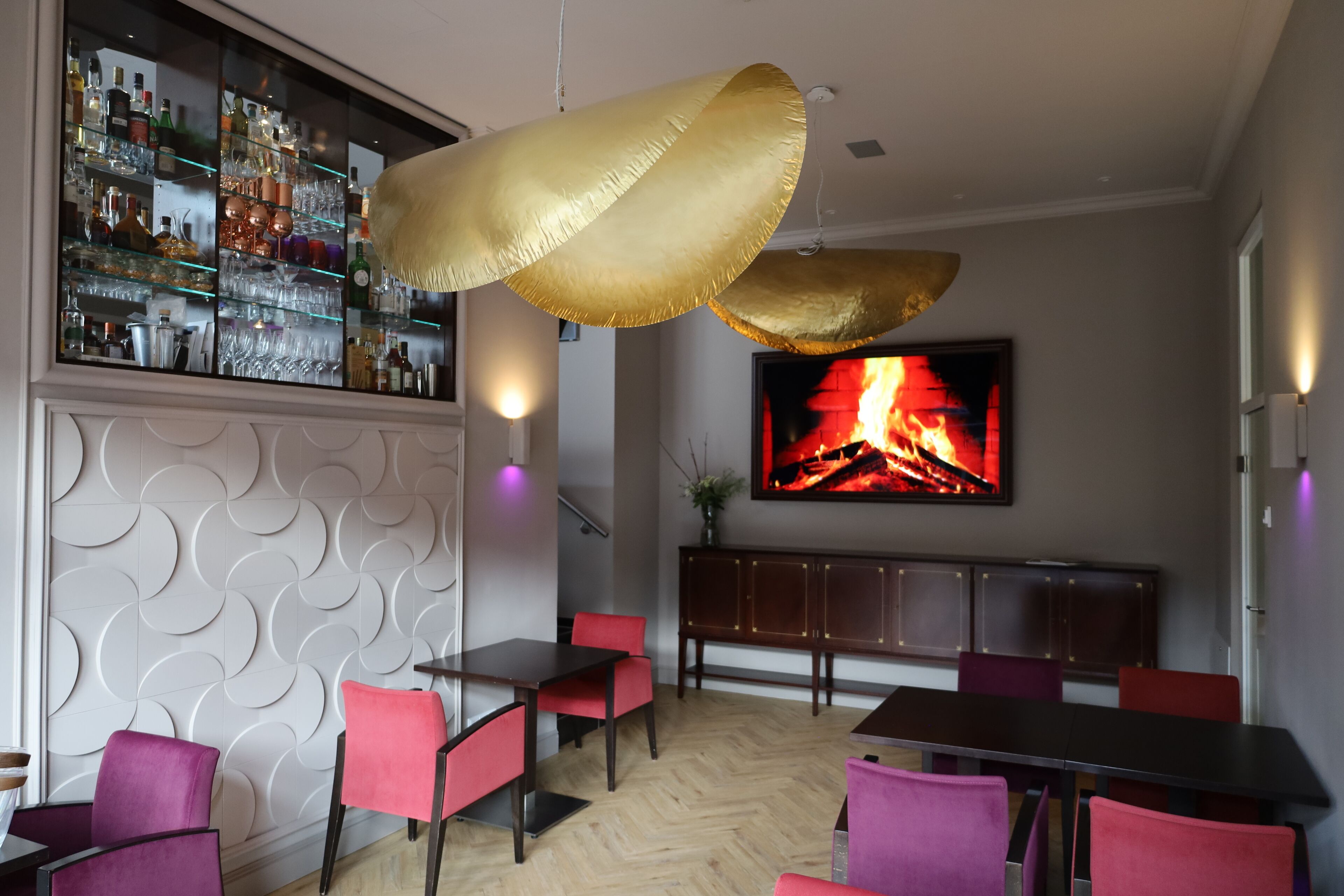 A modern bar area with a wine and liquor display, a large TV screen, and red and purple chairs.
