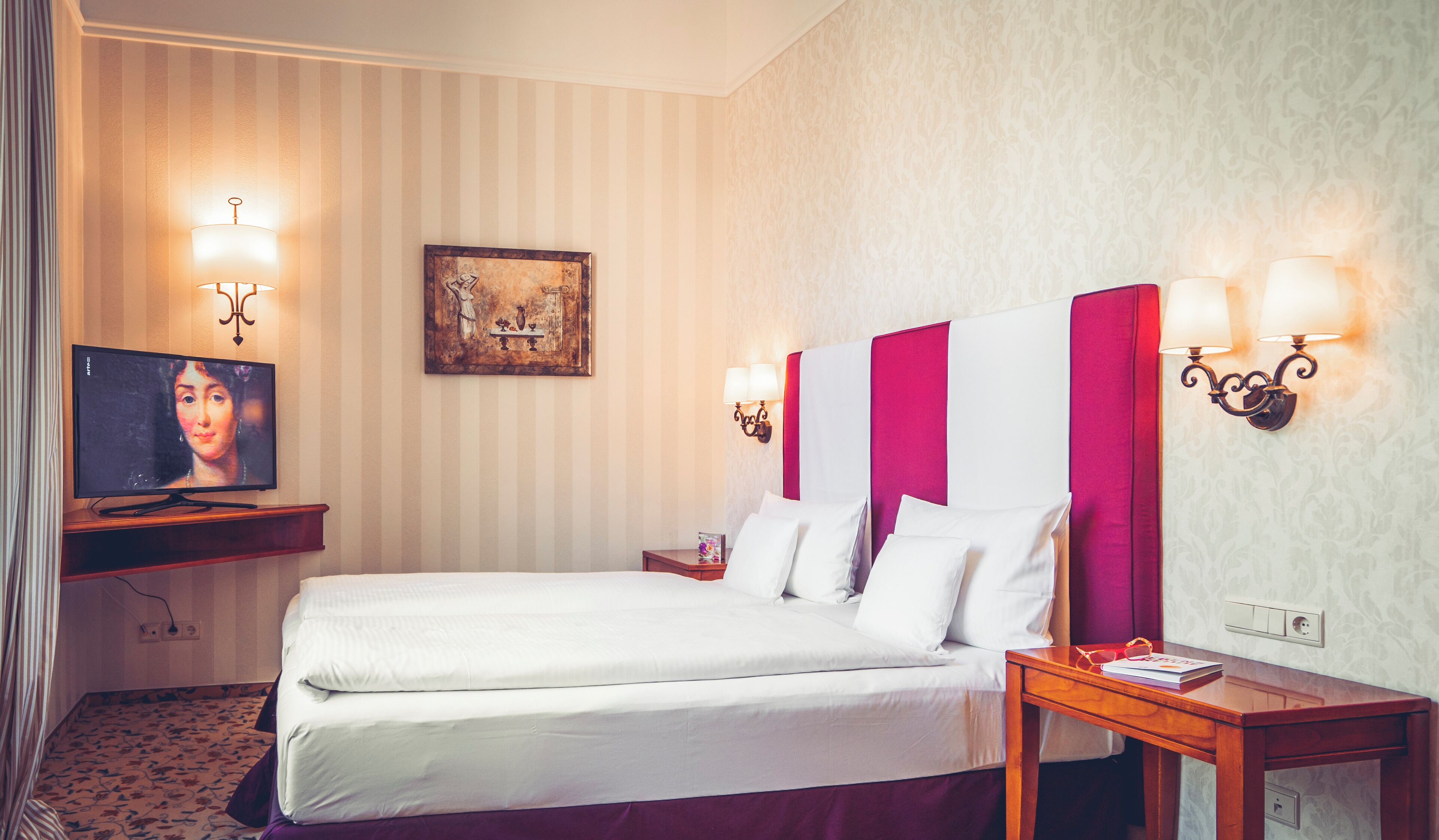 A hotel room with a bed, a television, a painting, and wall sconces.