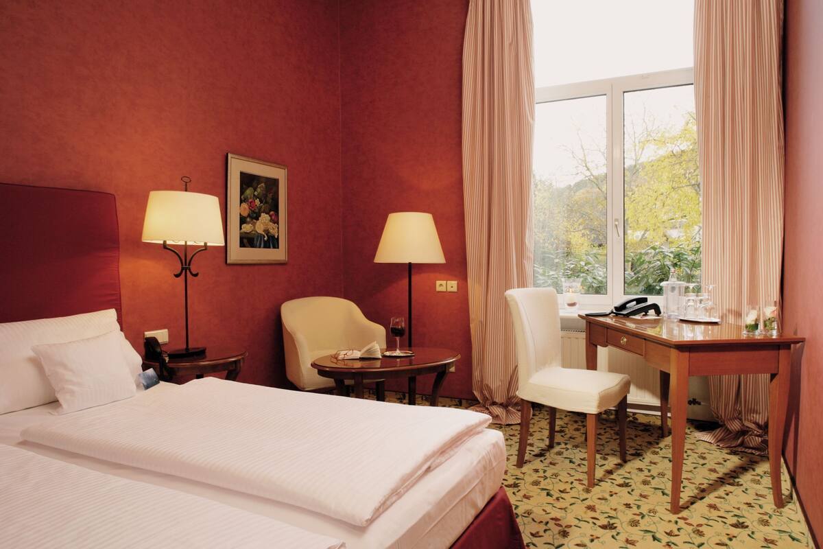A hotel room with a bed, two chairs, a desk, a lamp, a picture on the wall, and a window with curtains.