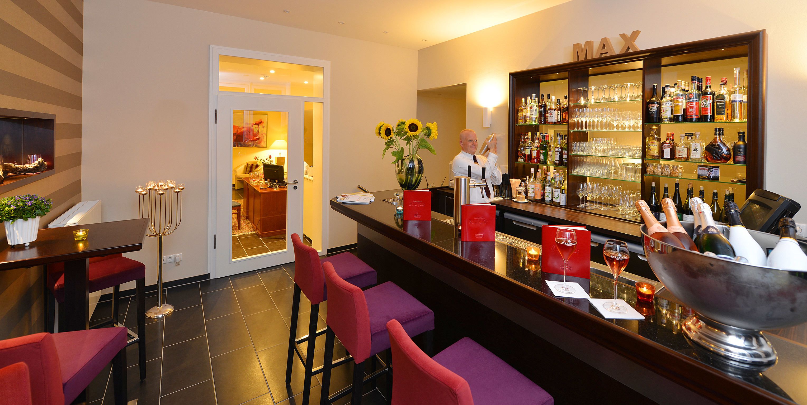 A bar area with a bar counter, red bar stools, a large wine bottle display, and a vase with sunflowers.