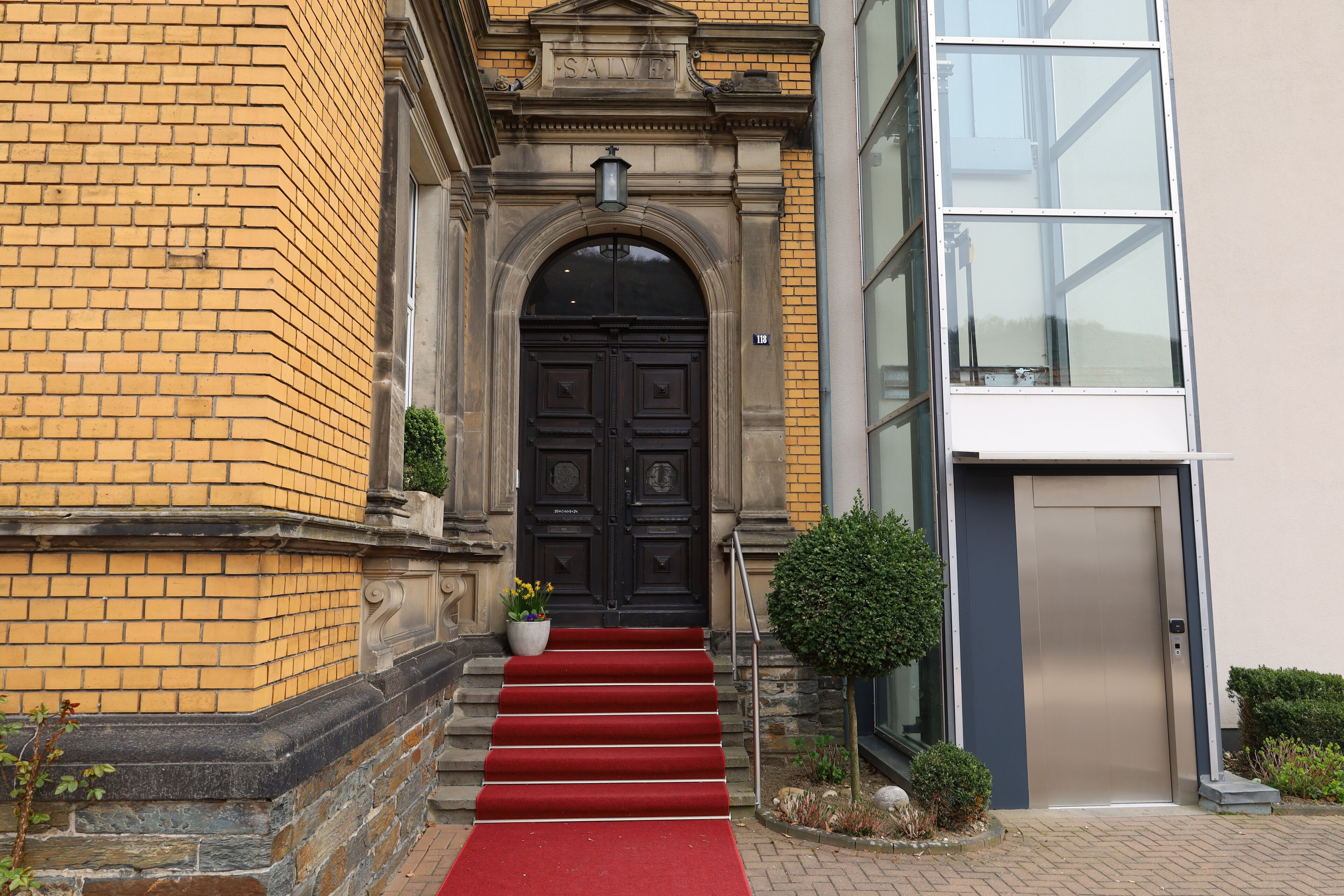 A building entrance with a red carpet leading to a black double door, a small potted plant, and a clear sky in the background.