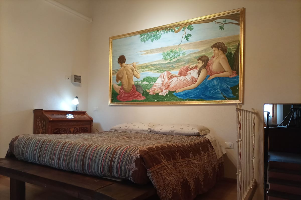 Un dormitorio con cama, banca, mesita de noche, un cuadro y un radiador.