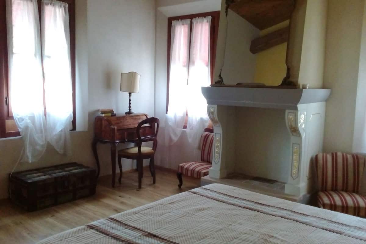 Un dormitorio con una cama de madera, un escritorio con lámpara, una silla, una mesita y una chimenea.