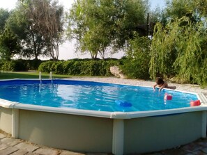 Ein überirdischer Swimmingpool mit umlaufender Holzterrasse, eingebettet in einen angelegten Garten mit Bäumen und Sträuchern.