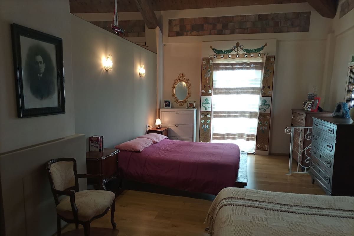 Un dormitorio con cama, una silla, cómoda, espejo y ventana con cortinas.
